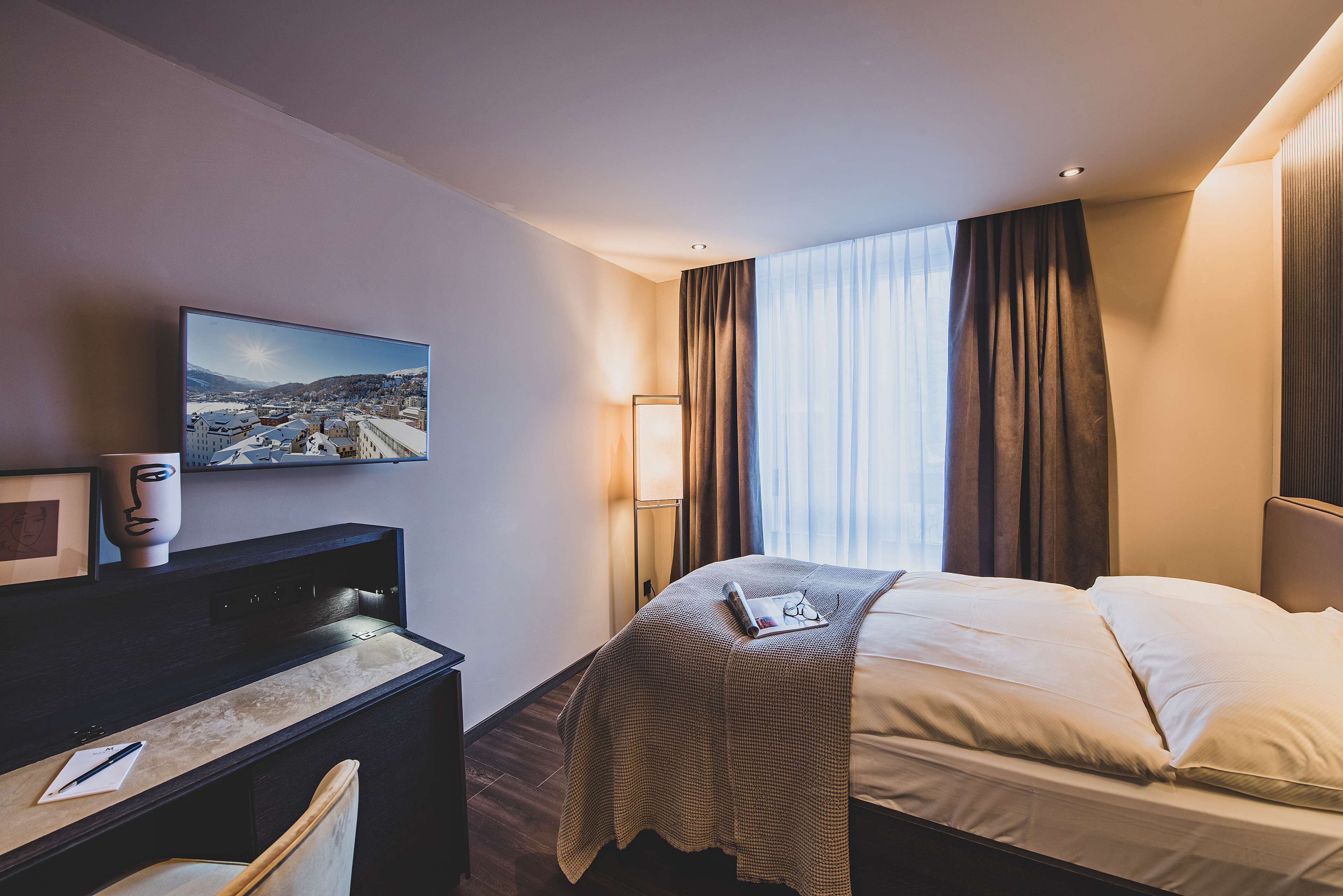 A hotel room with a bed, a desk, a chair, a lamp, and a painting on the wall.