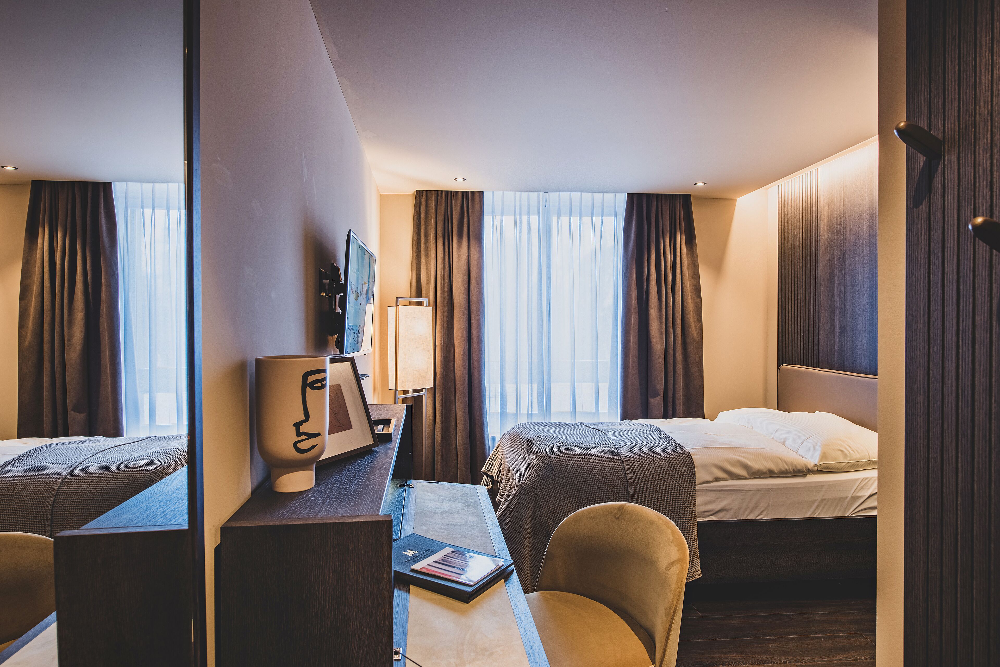 A hotel room with two beds, a desk with a lamp, a chair, and a view of the city through large windows.