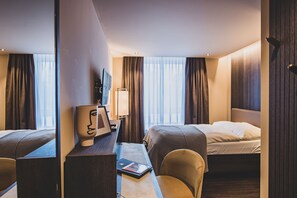 Habitación de hotel con dos camas, un escritorio con lámpara, una silla y vistas a la ciudad a través de amplias ventanas.