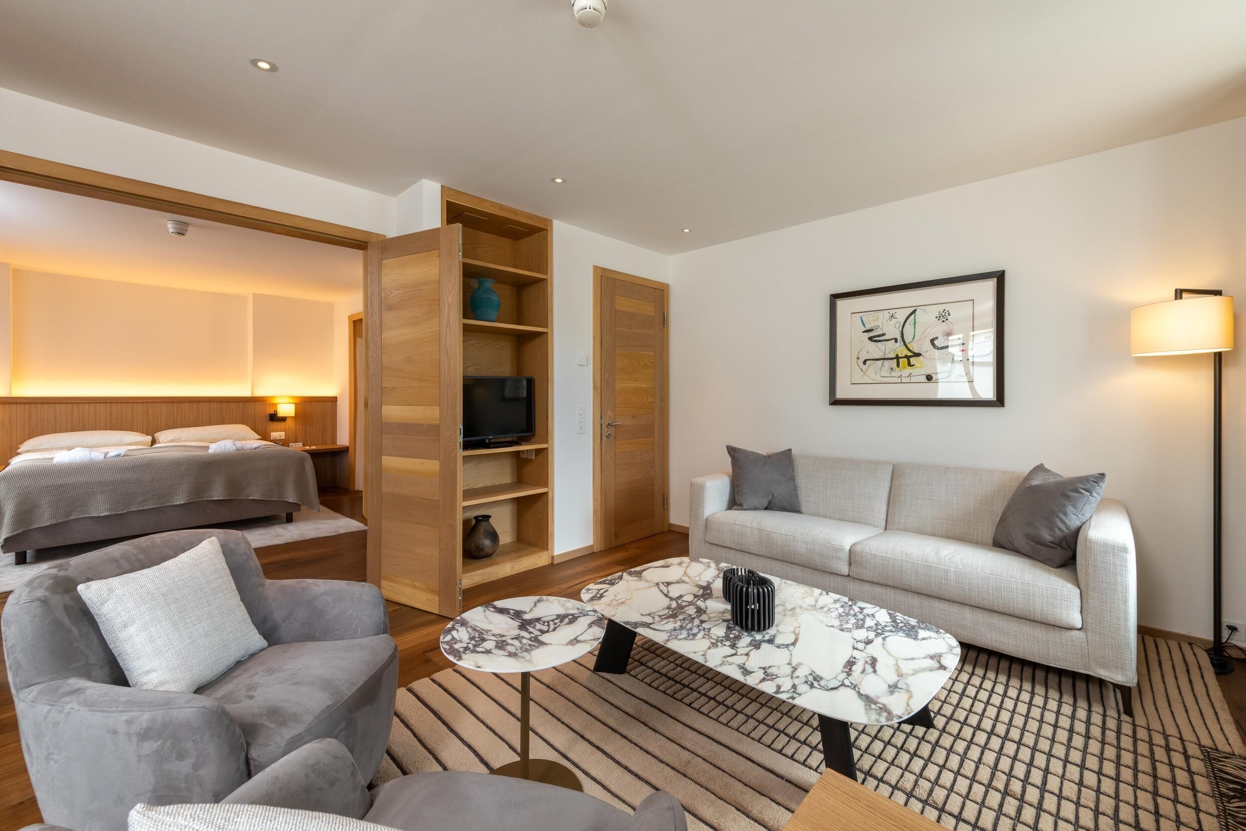 A modern hotel room with a bed, sofa, armchair, and a marble coffee table.