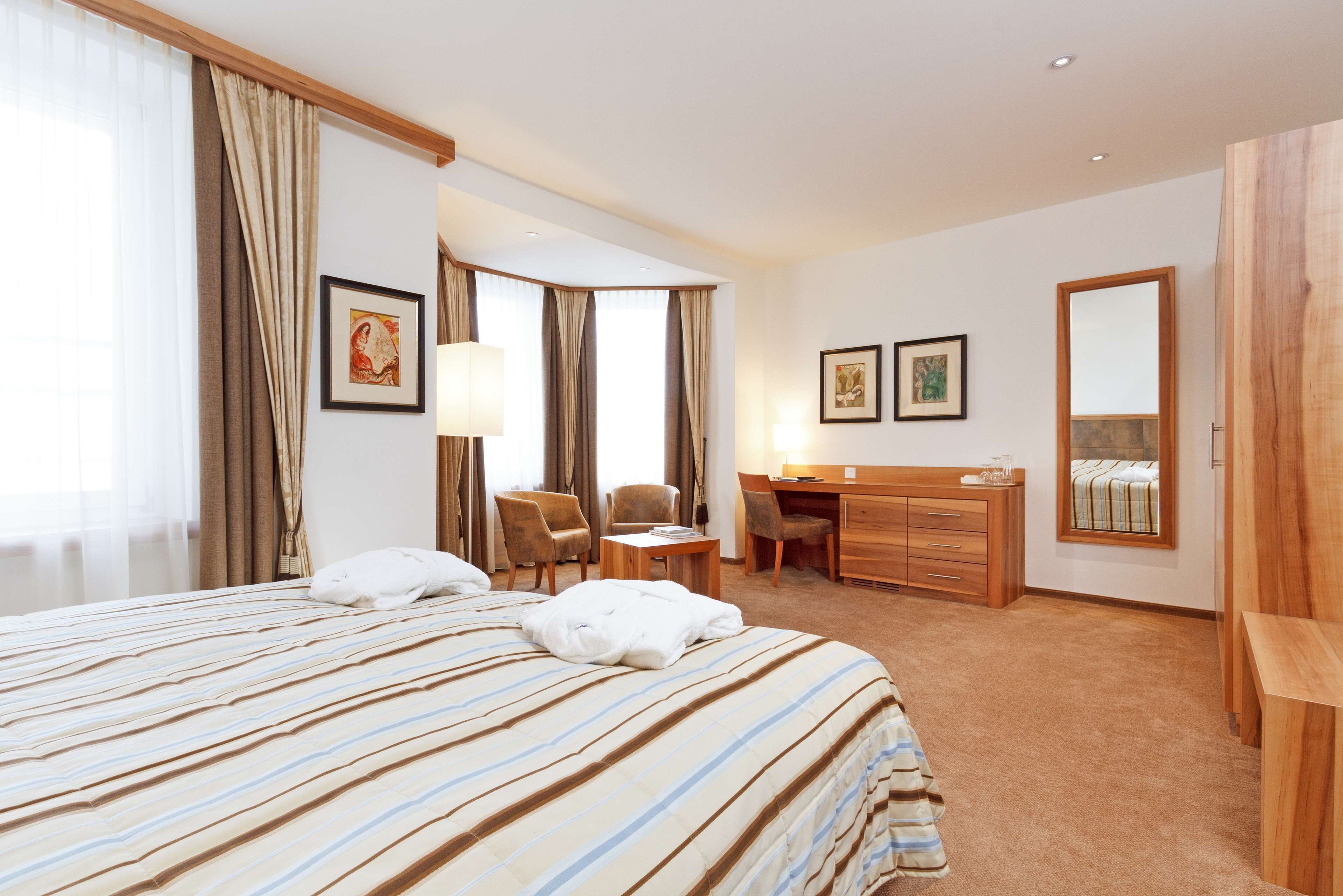 A hotel room with a large bed, a desk, two chairs, a dresser, and a mirror.