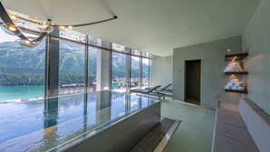 A modern indoor swimming pool with clear glass walls, lounge chairs, and a view of mountains and a lake. - Art Boutique Hotel Monopol (St. Moritz)