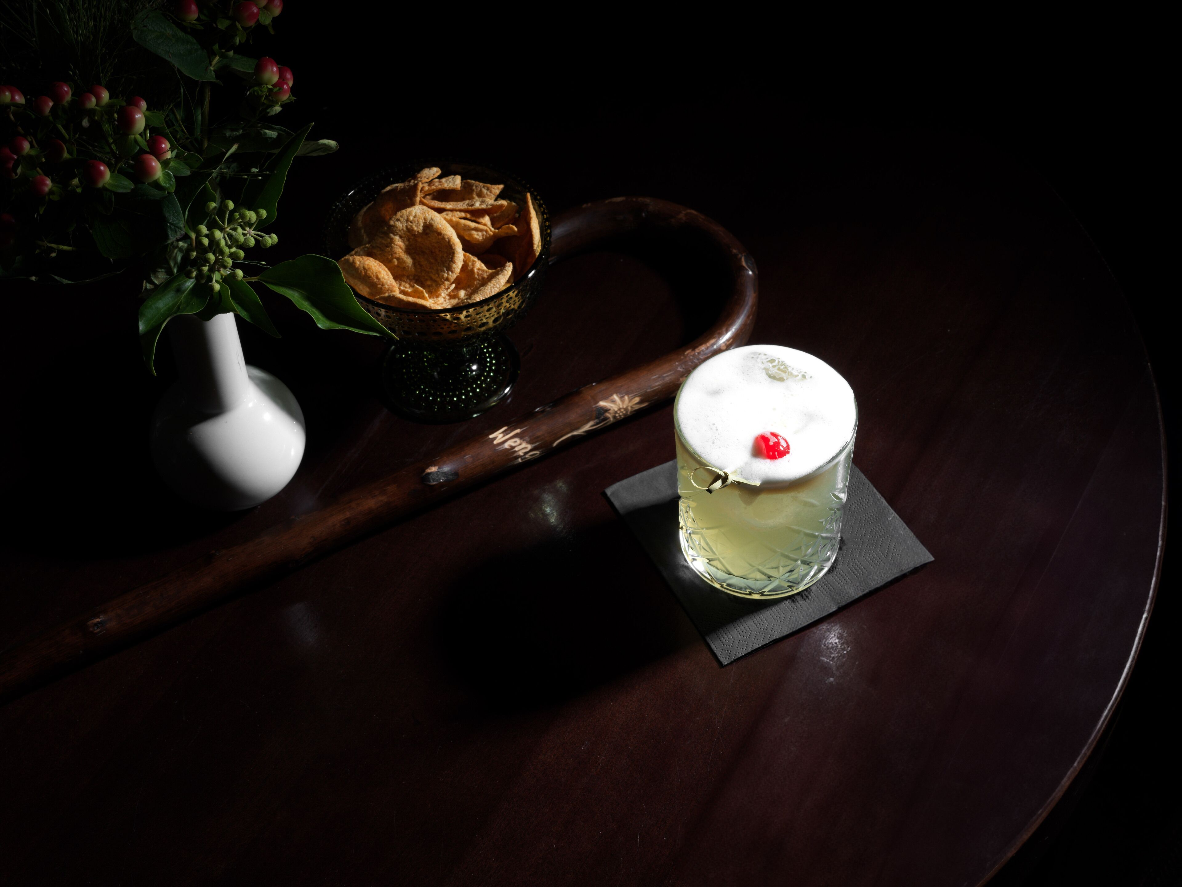A dark wooden table with a glass of a clear beverage topped with a cherry, a bowl of chips, and a white vase with green leaves.