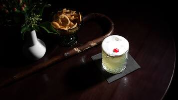 A dark wooden table with a glass of a clear beverage topped with a cherry, a bowl of chips, and a white vase with green leaves.