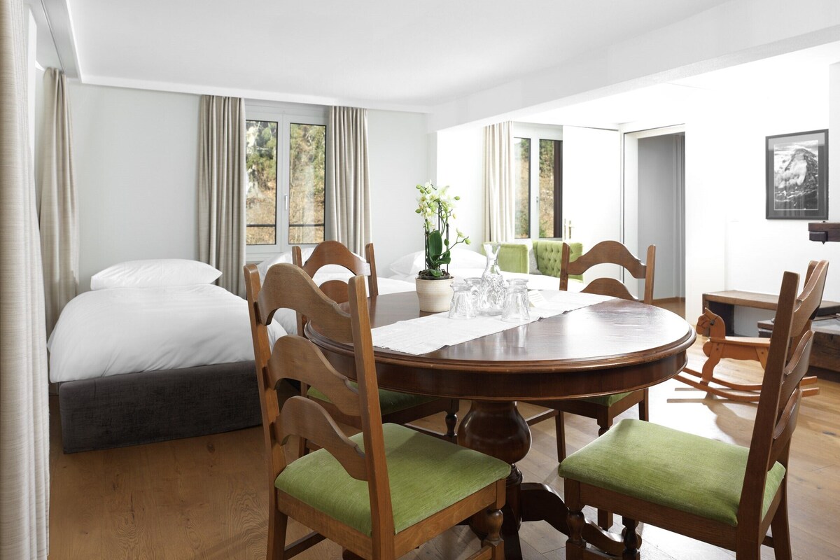 A room with a round wooden dining table surrounded by chairs with green cushions, a bed with white linens, and a window with curtains.