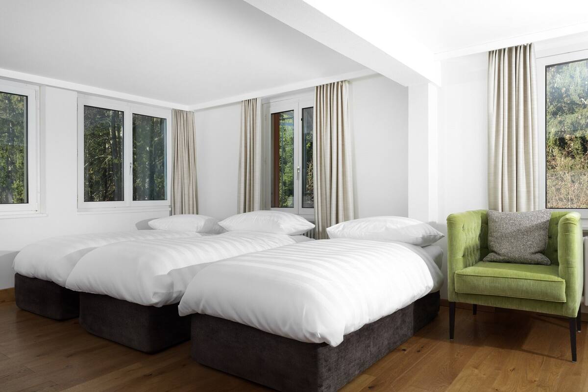 Ein modernes Schlafzimmer mit zwei großen Betten, einem grünen Sessel und großen Fenstern mit Vorhängen.