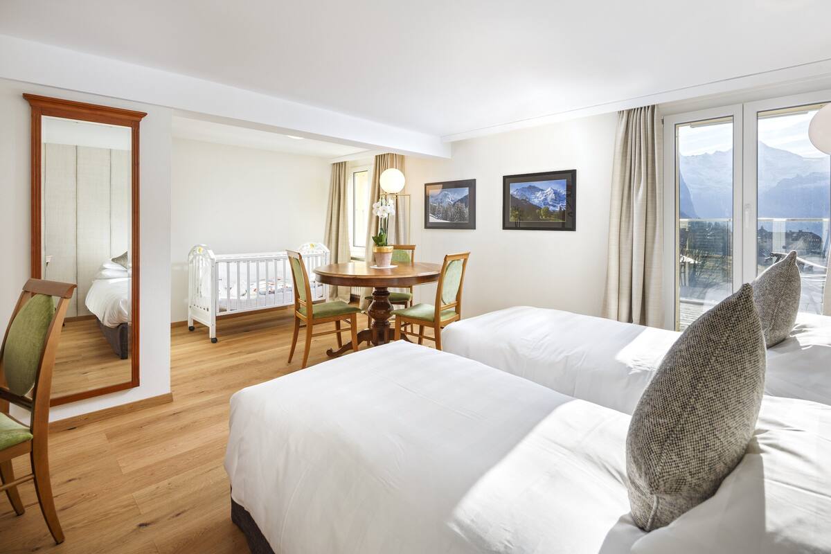 Ein Hotelzimmer mit zwei Betten, einem Esstisch mit Stühlen, einem kleinen Balkon und Blick auf die Berge.