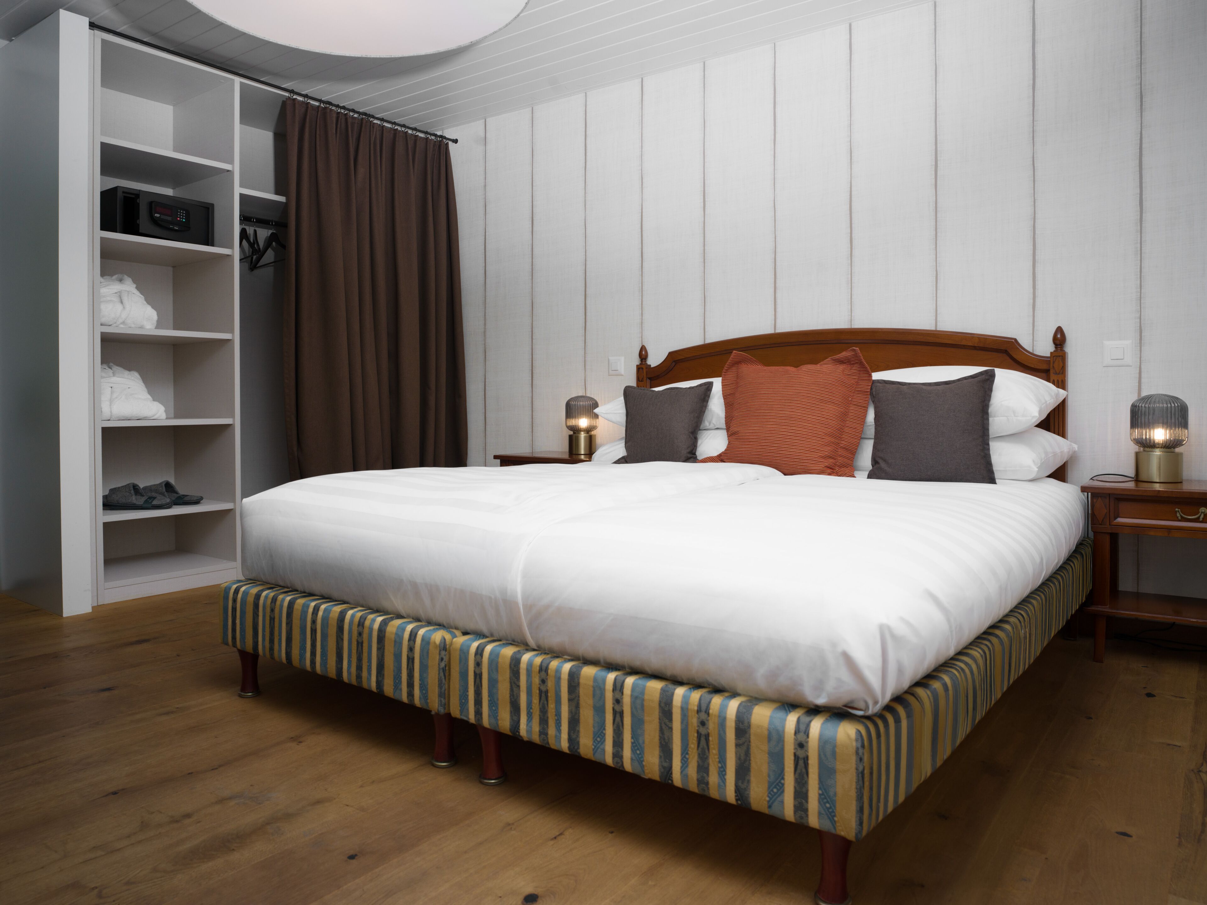 Una cama bien hecha con cabecera de madera, una mesita de noche con lámpara y un amplio armario con cortina.