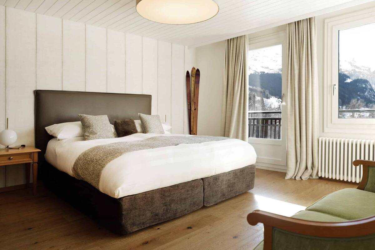 Ein Schlafzimmer mit einem großen Bett, einem hölzernen Nachttisch, einem grünen Sessel und Bergblick durch ein Fenster.