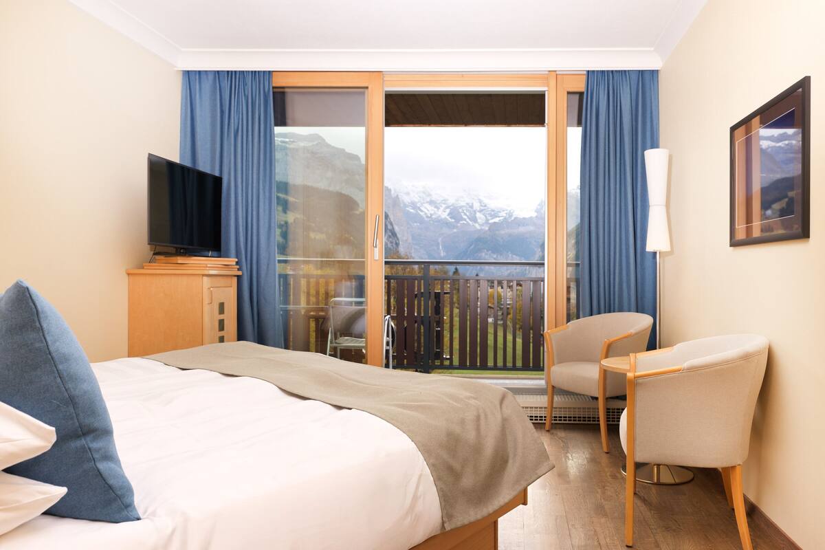 Ein Hotelzimmer mit Bett, Fernseher, Stuhl und Balkon mit Bergblick.