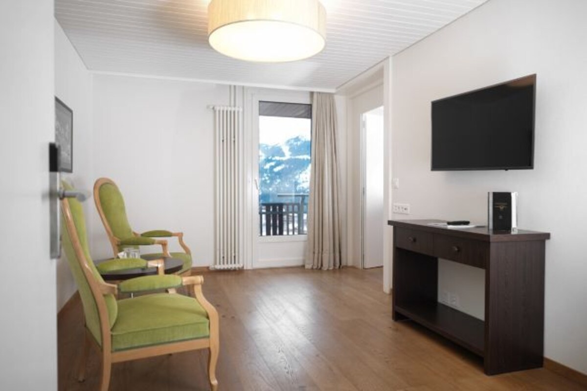 Ein Zimmer mit Holzboden, einem grünen Sessel, einem dunklen Holztisch, einem Flachbildfernseher und Bergblick durch ein Fenster.