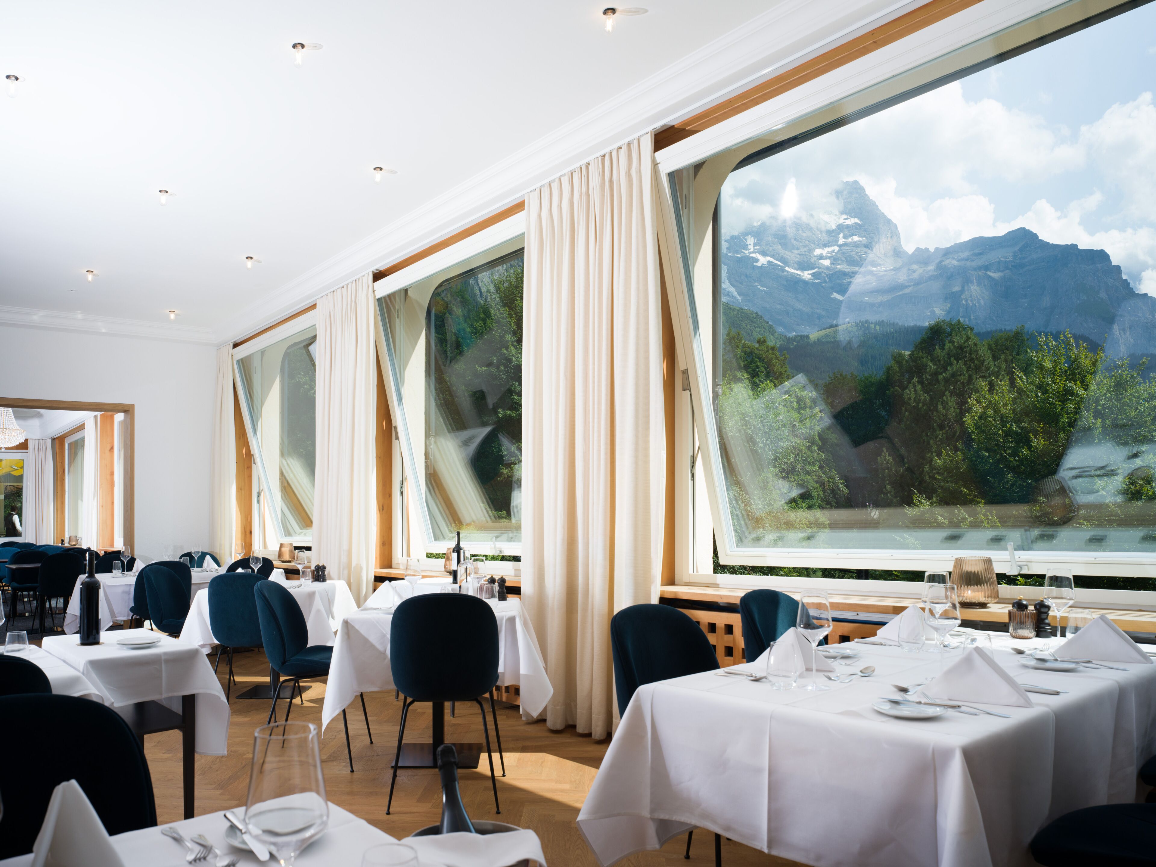 Restaurante com mesas preparadas para refeições, janelas amplas com cortinas e vista para montanhas.