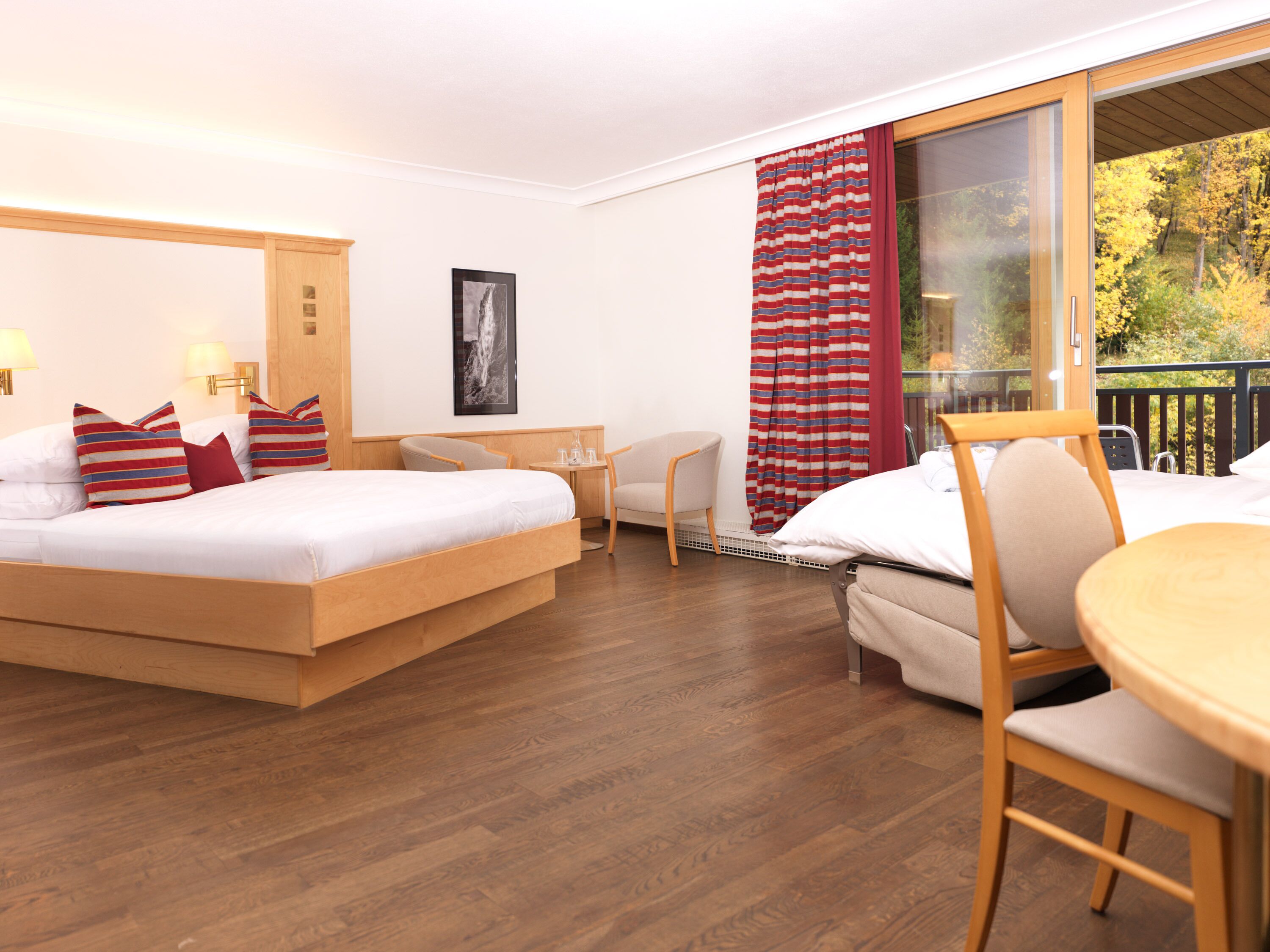 A hotel room with a bed, a desk, a chair, and a balcony with a view of trees.