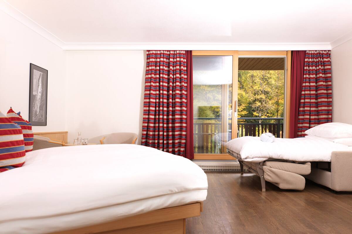Une chambre d’hôtel avec un lit, une chaise, une petite table et un balcon agrémenté de rideaux rouges.