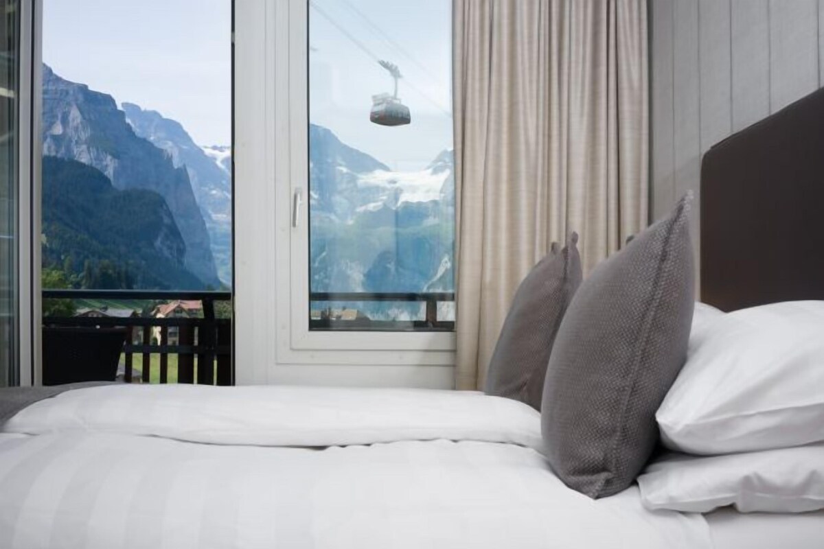 Ein Bett mit weißer Bettwäsche und grauen Kissen, ein Fenster mit Bergblick und ein teilweise gezogener Vorhang.