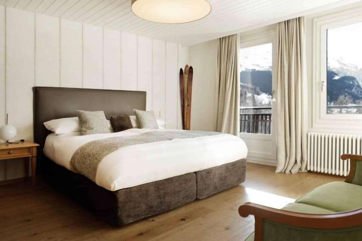 Un dormitorio con una cama grande, una mesita de noche, una silla, una ventana con cortinas y vistas a las montañas.