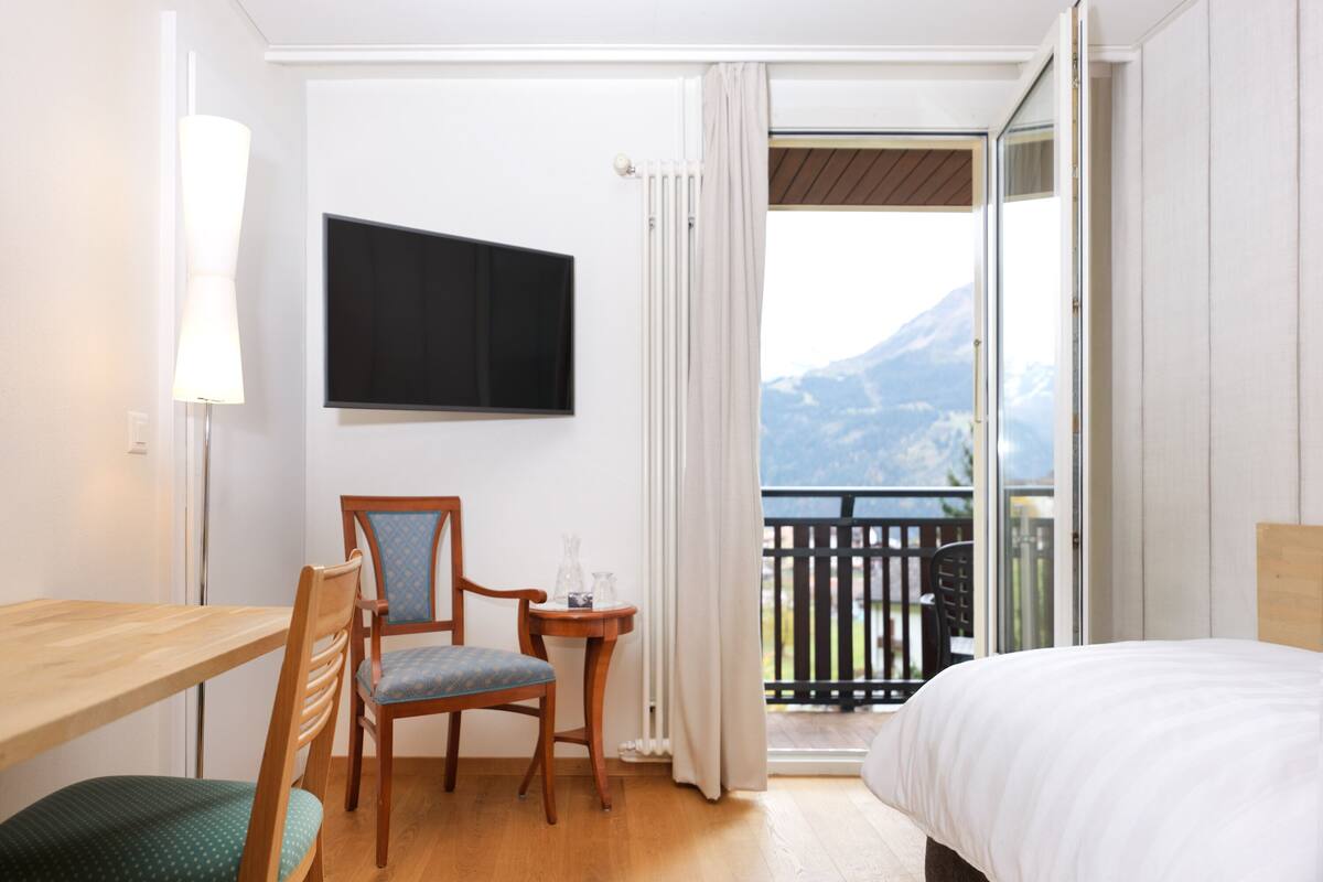 Ein Hotelzimmer mit einem Holztisch, einem Stuhl, einem Bett, einem Fernseher und einem Balkon mit Bergblick.