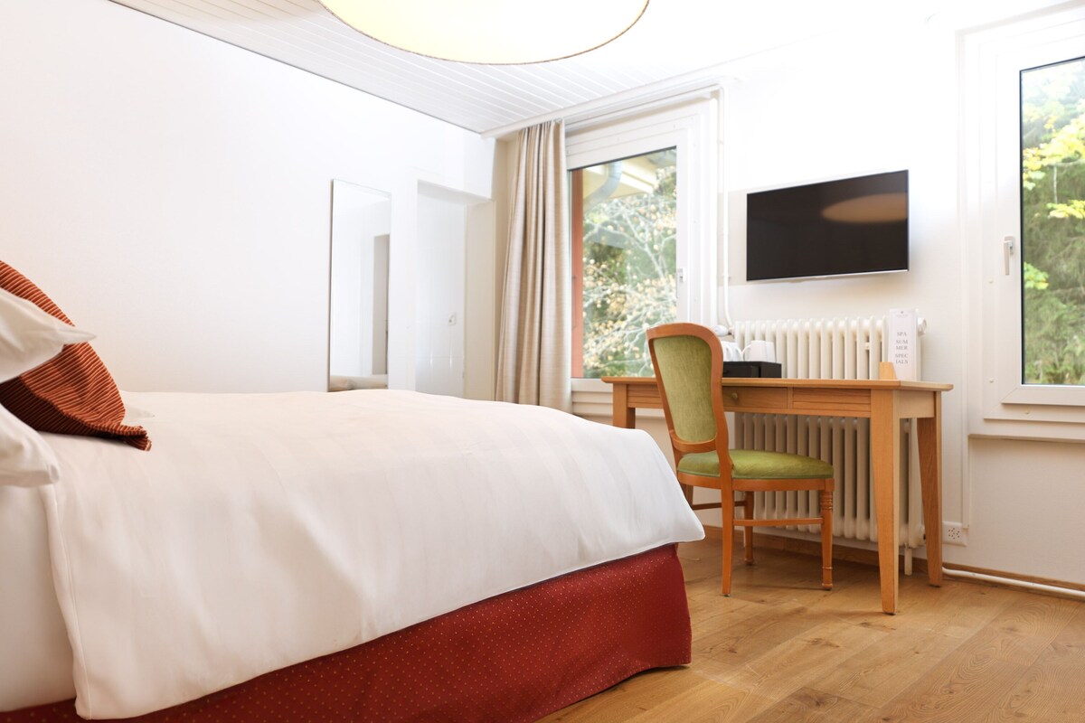 A hotel room with a bed, a desk, a chair, a television, and a window with a view of greenery.