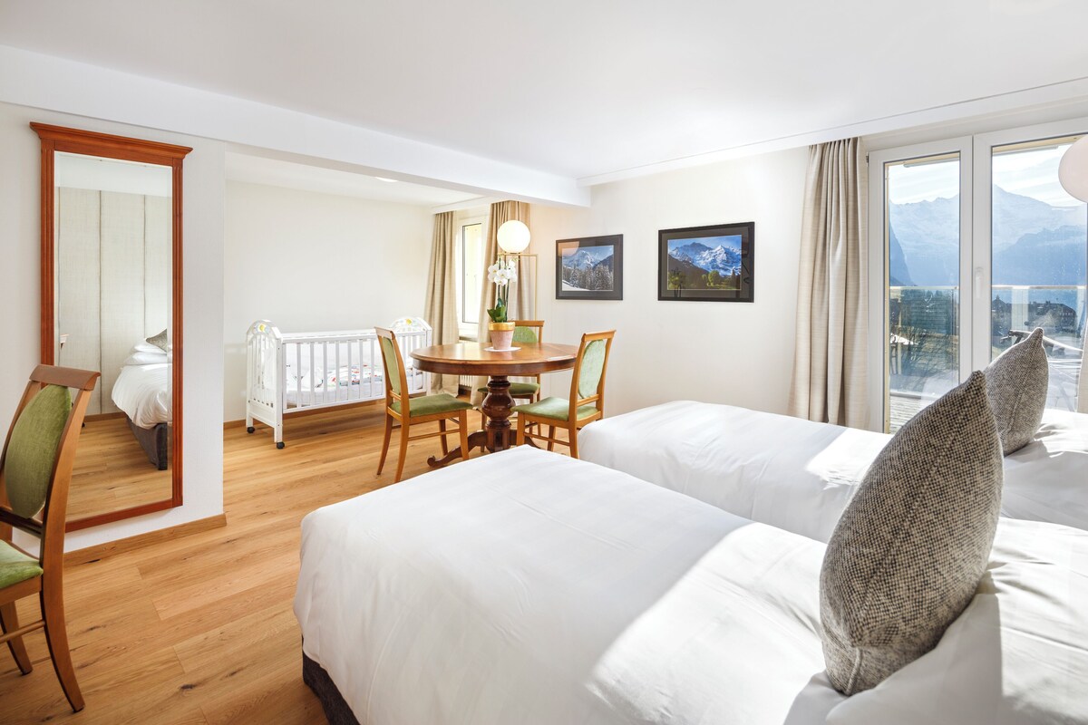 A hotel room with two beds, a dining table with chairs, a small balcony, and mountain views.