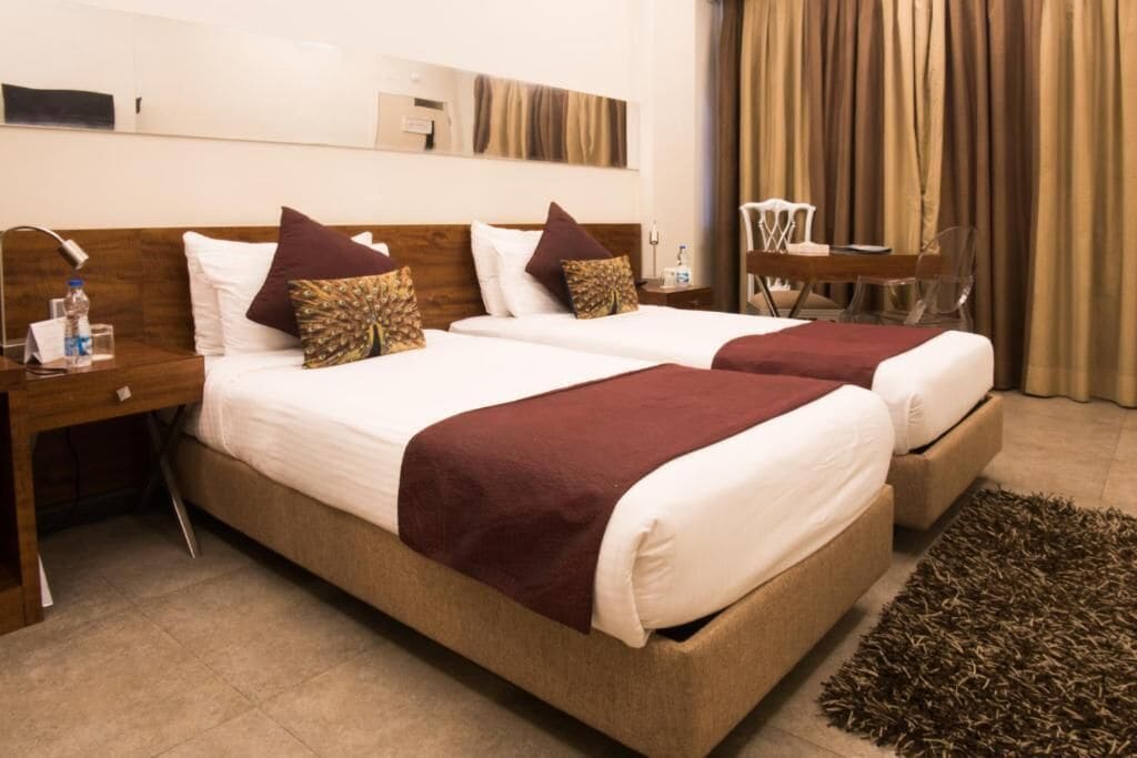 Une chambre d’hôtel avec deux lits, une tête de lit en bois, une table de chevet avec une lampe, une petite table avec des chaises et une grande fenêtre avec des rideaux.