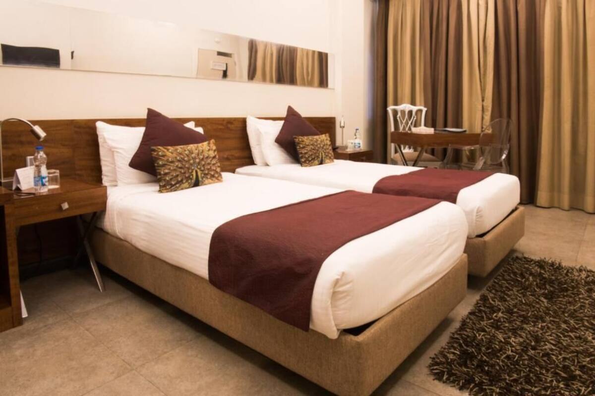 A hotel room with two beds, a wooden headboard, a bedside table with a lamp, a small table with chairs, and a large window with curtains.