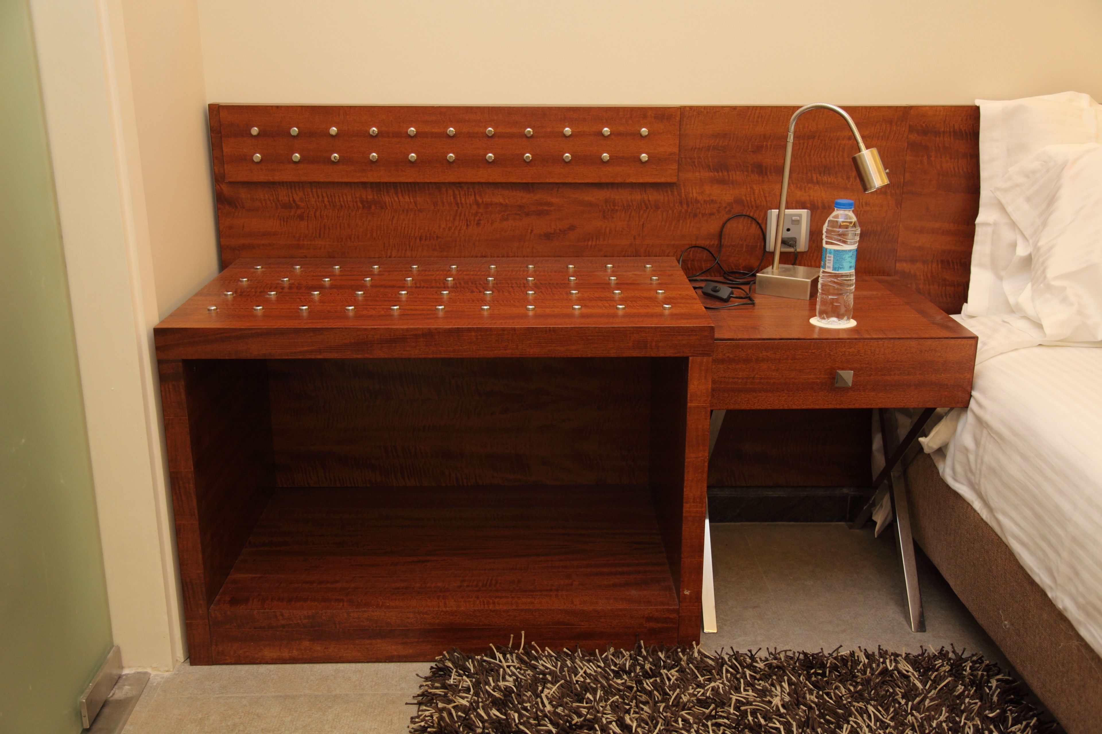 A wooden nightstand with a water bottle and a lamp, next to a bed with white bedding.