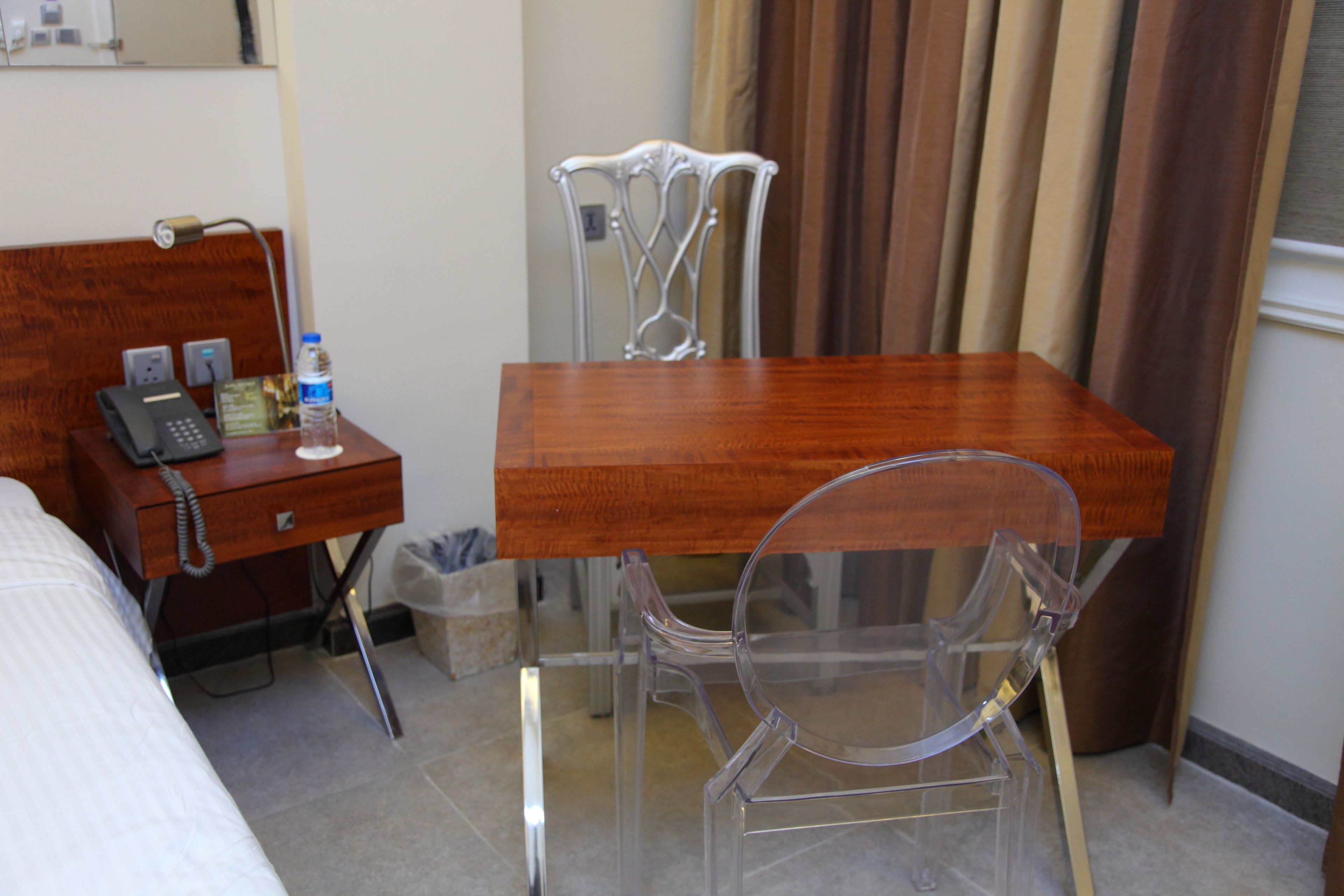 A hotel room with a wooden desk, a clear plastic chair, a telephone, and a trash bin.