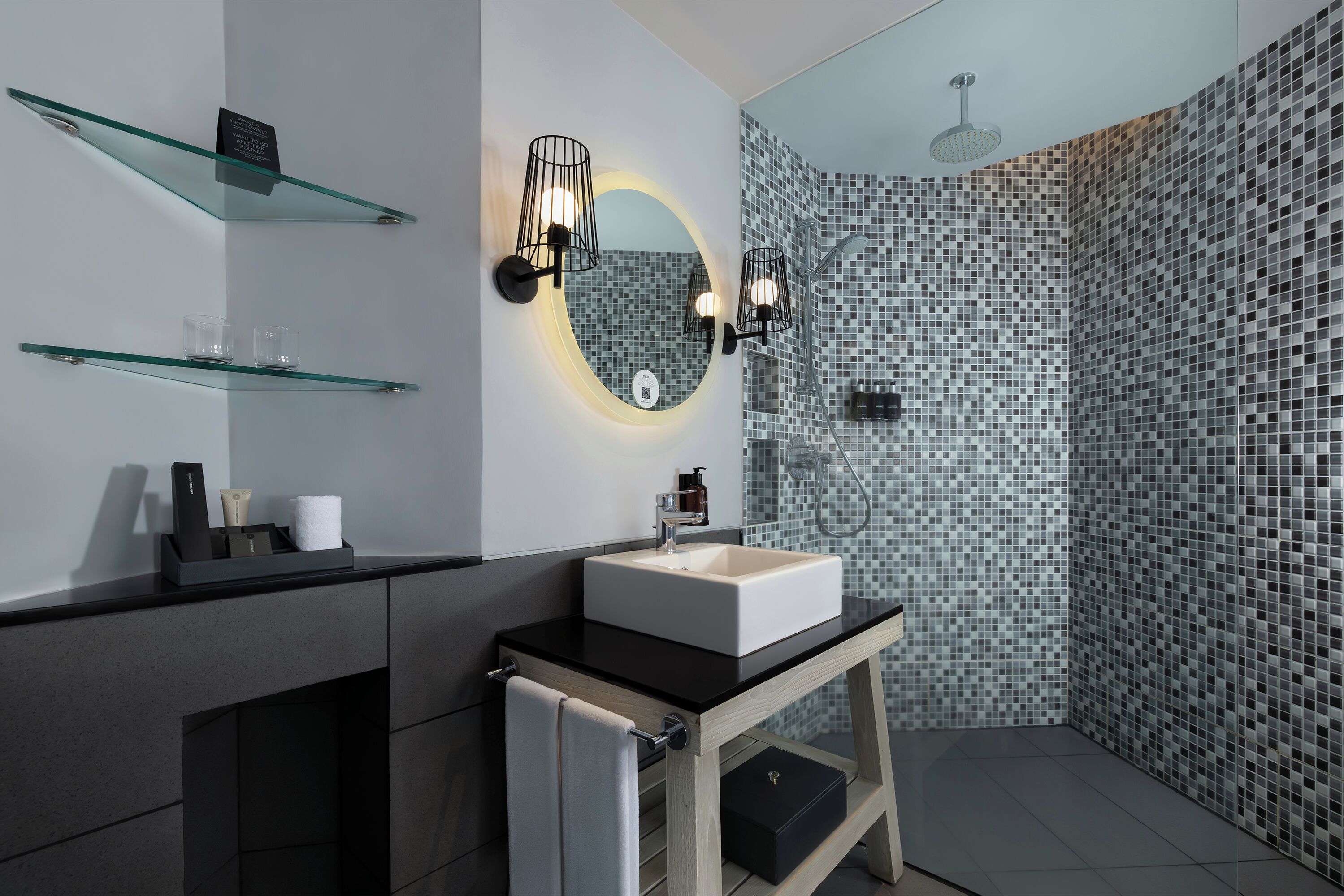 Un baño moderno con un estante de vidrio, un espejo redondo, una encimera negra y una zona de ducha con pared de azulejos.