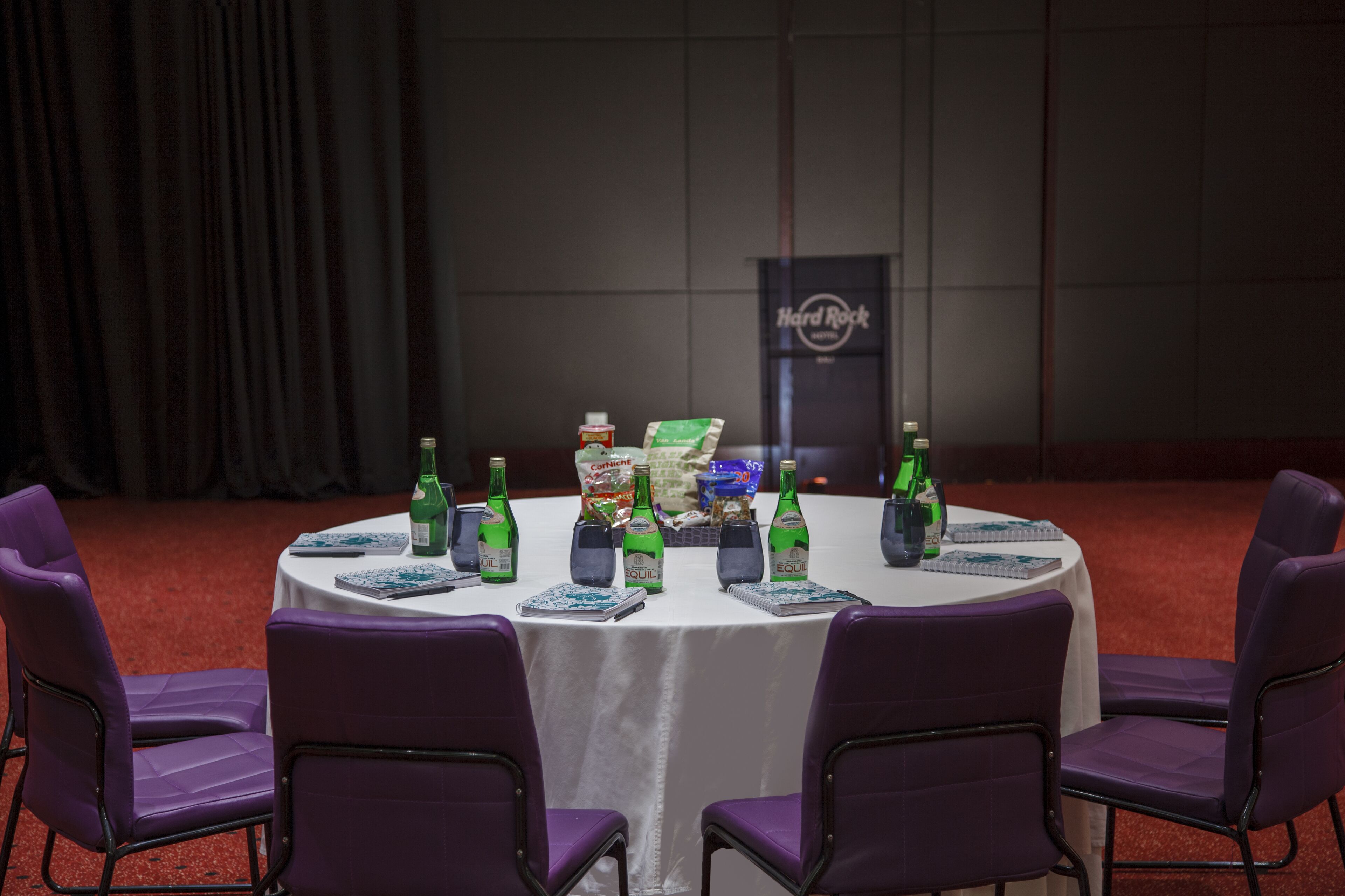 Ein runder Tisch mit violetten Stühlen, einer weißen Tischdecke und verschiedenen Gegenständen, darunter Flaschen, Snacks sowie ein Hard Rock Schild im Hintergrund.
