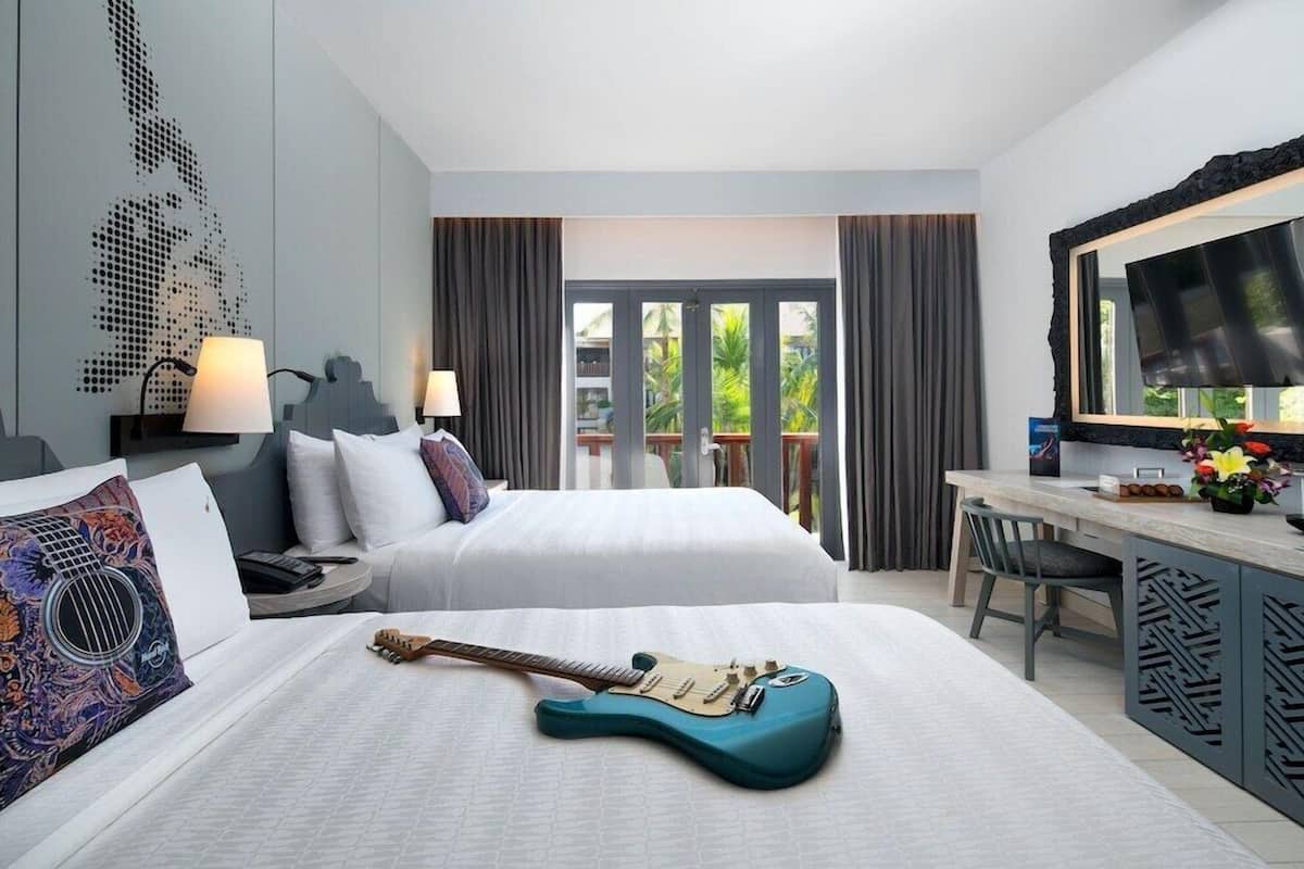 A hotel room with a bed, a desk, a guitar, a TV, and a balcony with curtains.