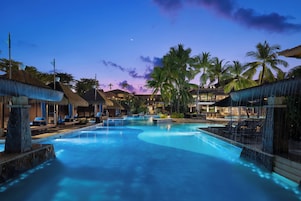 A large outdoor swimming pool surrounded by palm trees and thatched-roof structures.