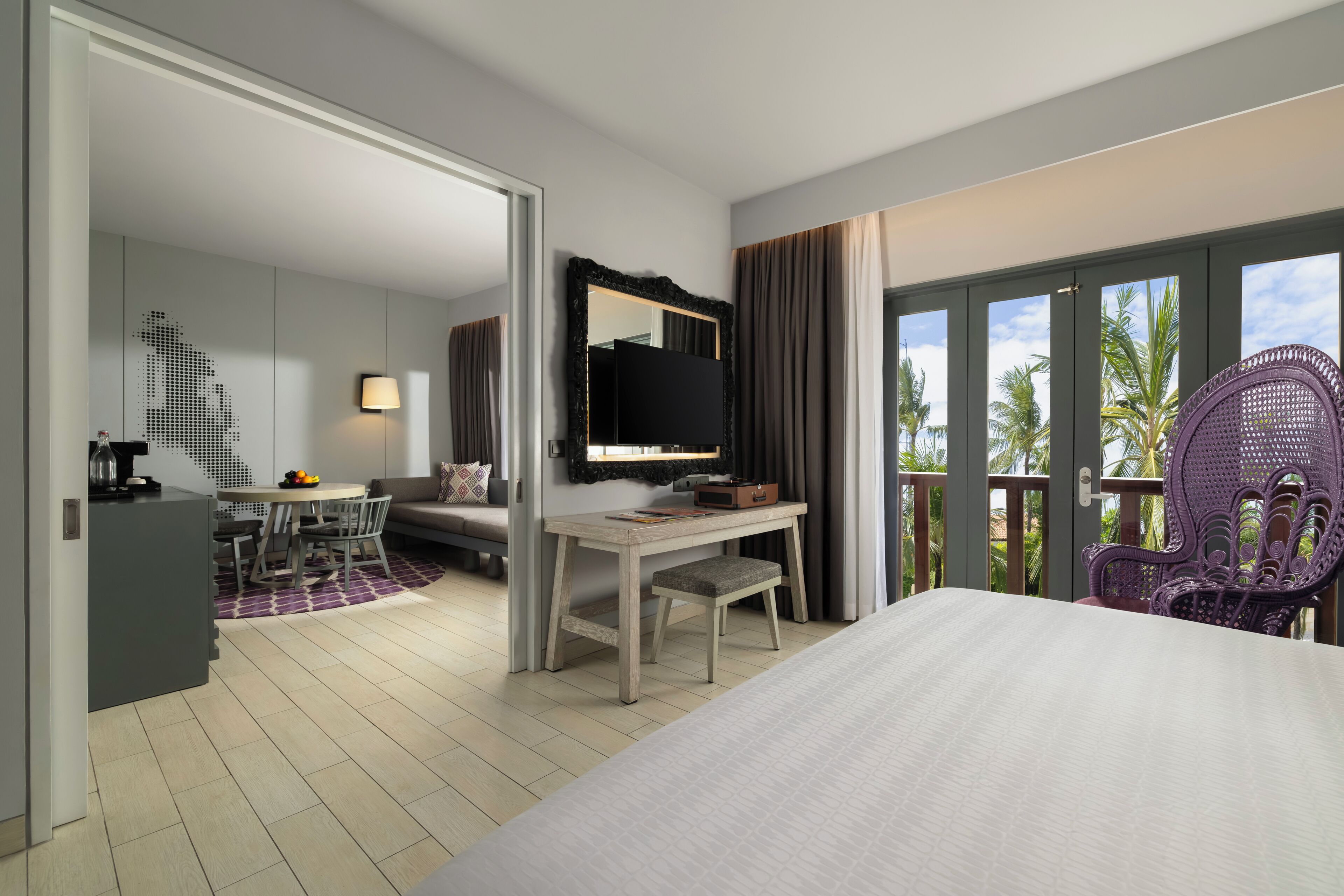A hotel room with a bed, a desk, a television, a chair, and a balcony with a view of palm trees.