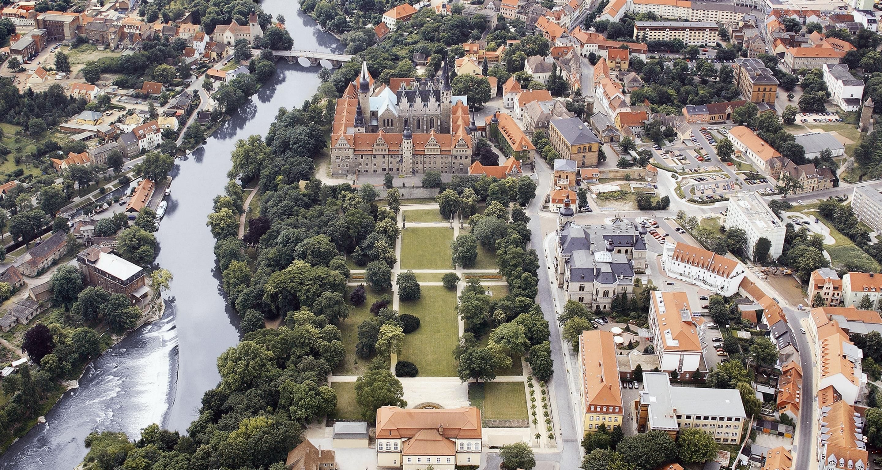 Vista aérea de un pueblo histórico con un río, edificios y una imponente construcción similar a un castillo.