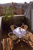 Una terraza de madera con una mesa y sillas, plantas en macetas y vistas de edificios al fondo.