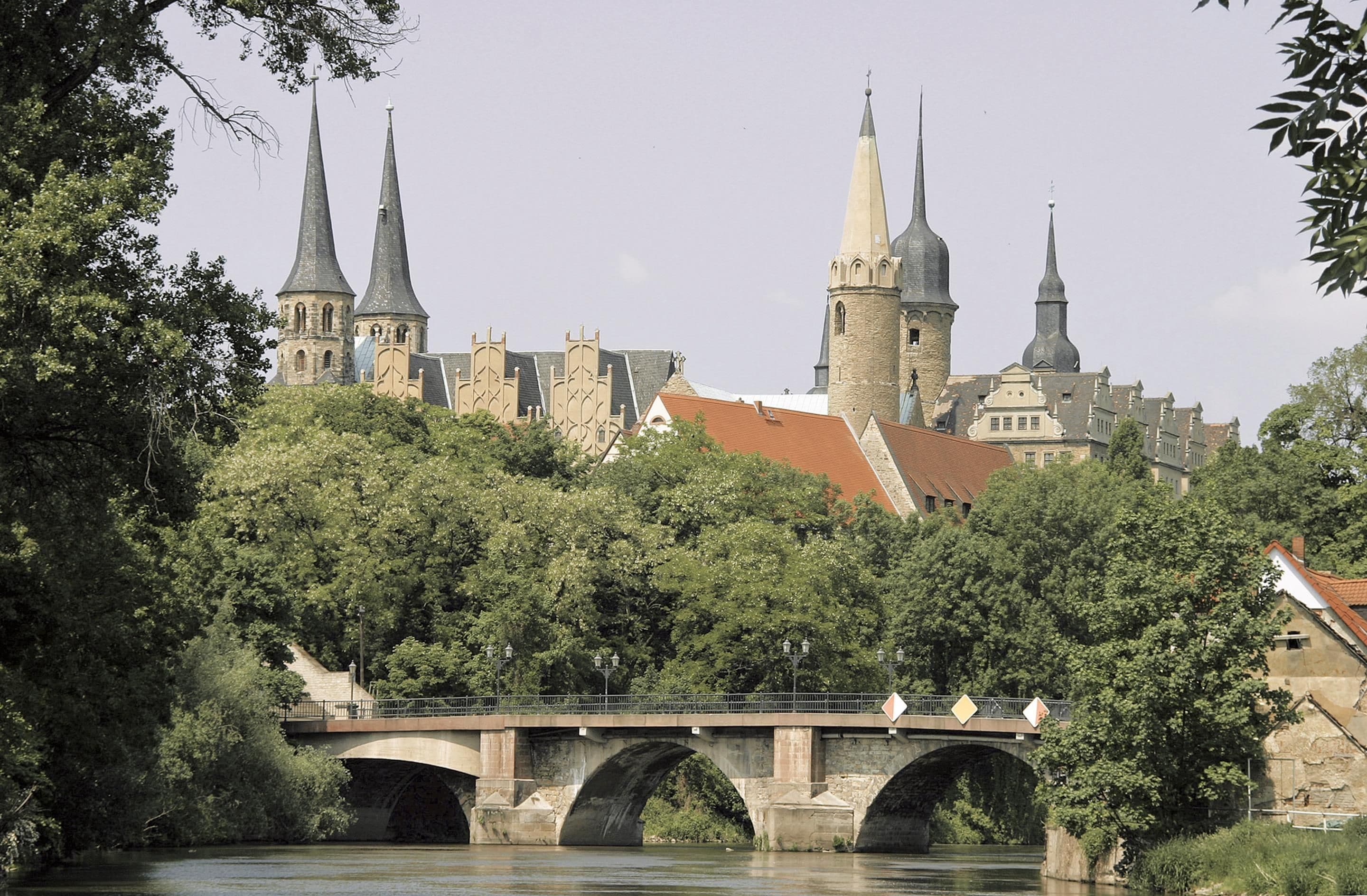 A stone bridge over a river with a town in the background featuring historic buildings and spires.