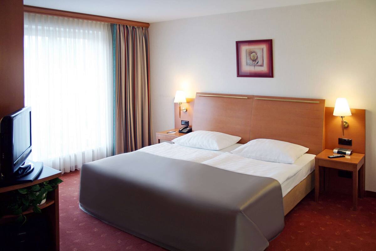 Habitación de hotel con una cama grande, dos mesitas de noche con lámparas, un televisor y una ventana con cortinas.
