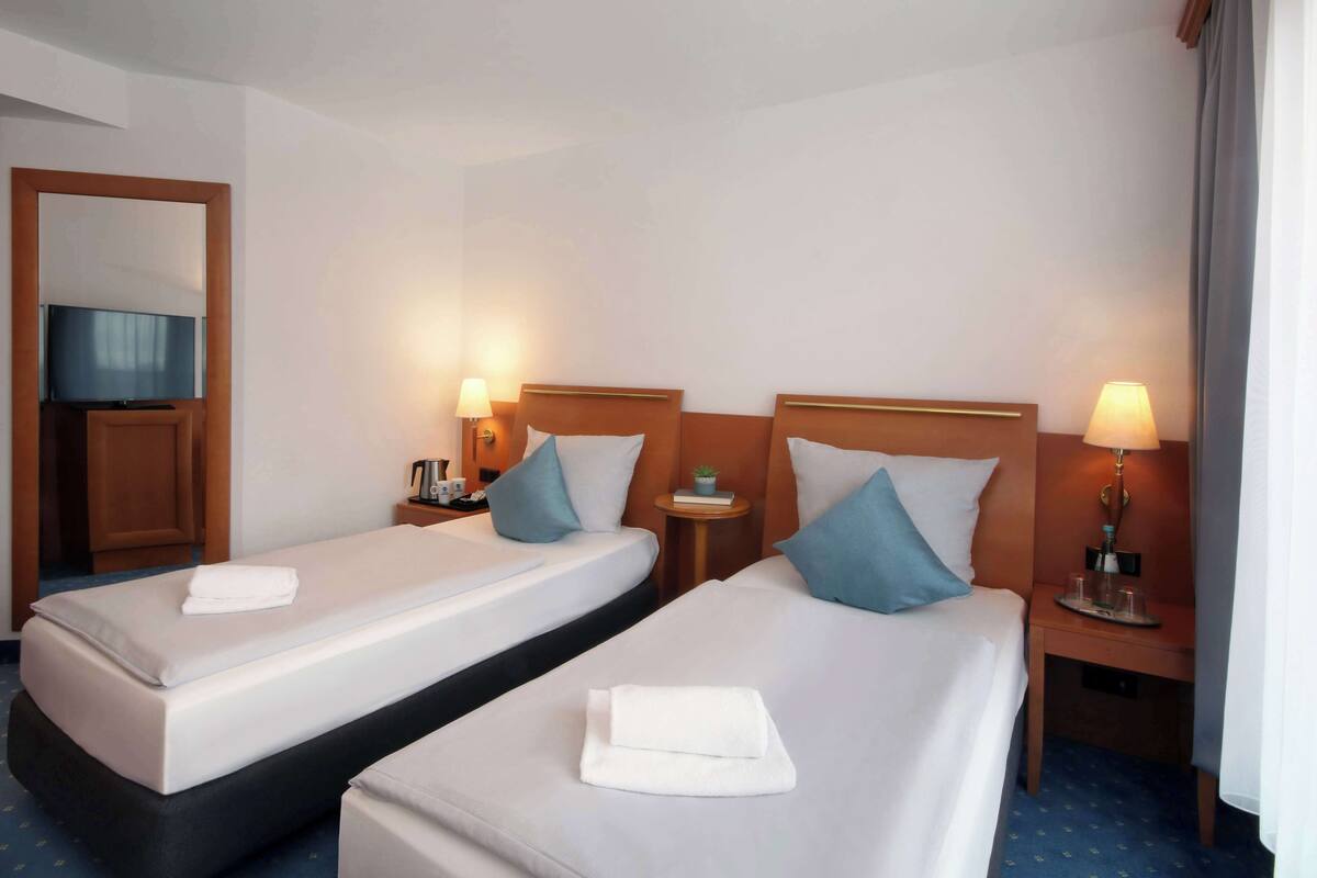 Habitación de hotel con dos camas, cada una con una almohada y una toalla doblada. Hay mesitas de noche con lámparas y una planta pequeña sobre una de ellas.