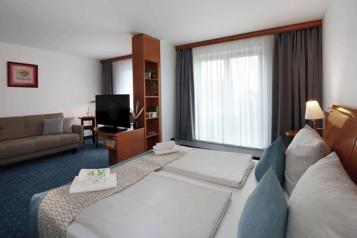 Habitación de hotel con cama, sofá, televisión, estantería y ventana con cortinas.
