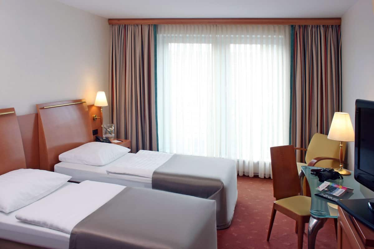 Una habitación de hotel con dos camas, un escritorio con silla, televisión y ventana con cortinas.