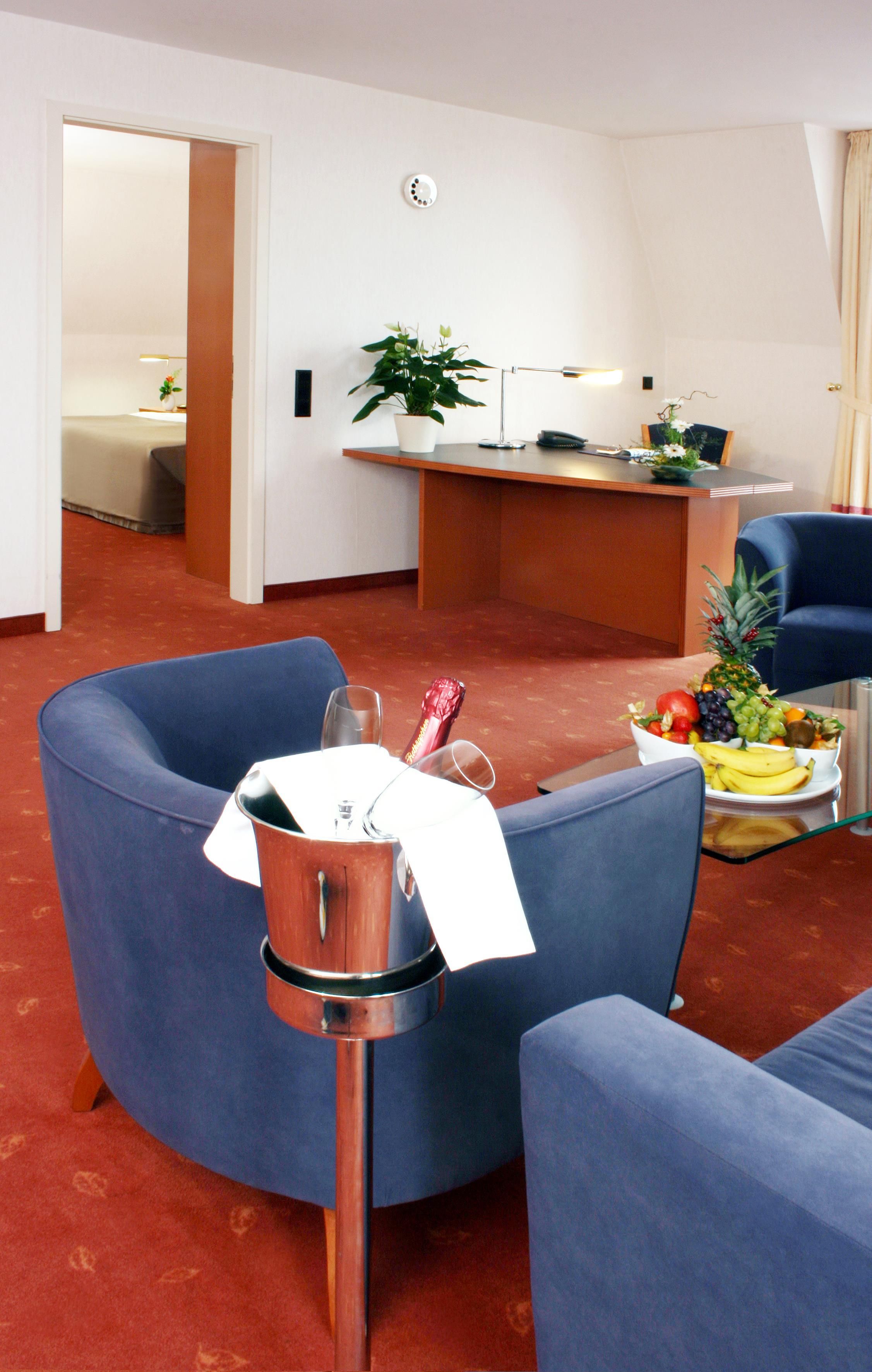 A hotel room with a blue armchair, a small table with a fruit bowl, a desk with a plant, and a bed in the background.