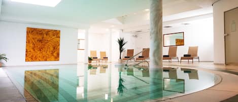 A modern indoor swimming pool with clear water, surrounded by lounge chairs and a large abstract painting.