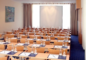 Een vergaderzaal met rijen tafels, stoelen en waterflessen, een projectiescherm en een schilderij aan de muur.
