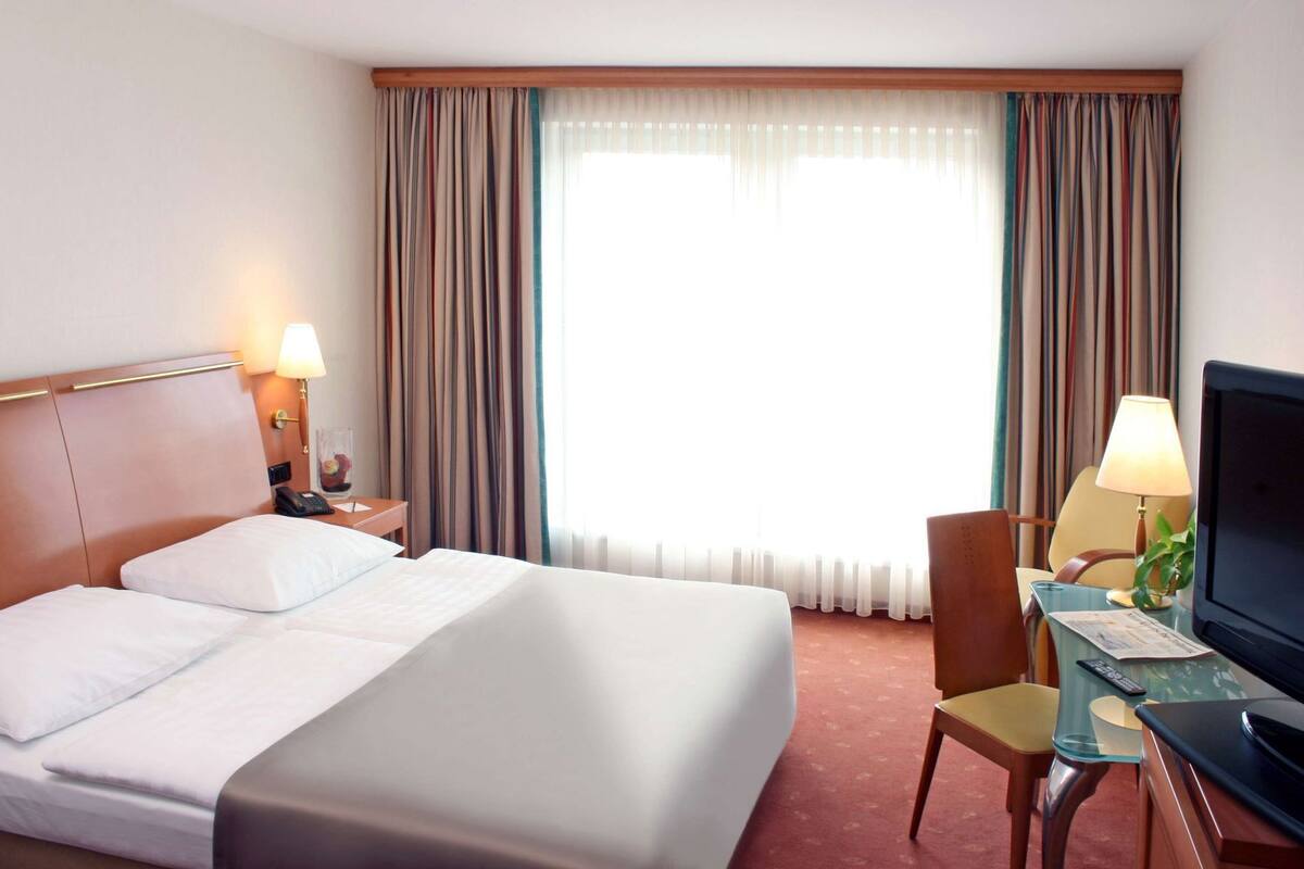 Una habitación de hotel con una cama, un escritorio con silla, un televisor y una ventana con cortinas.
