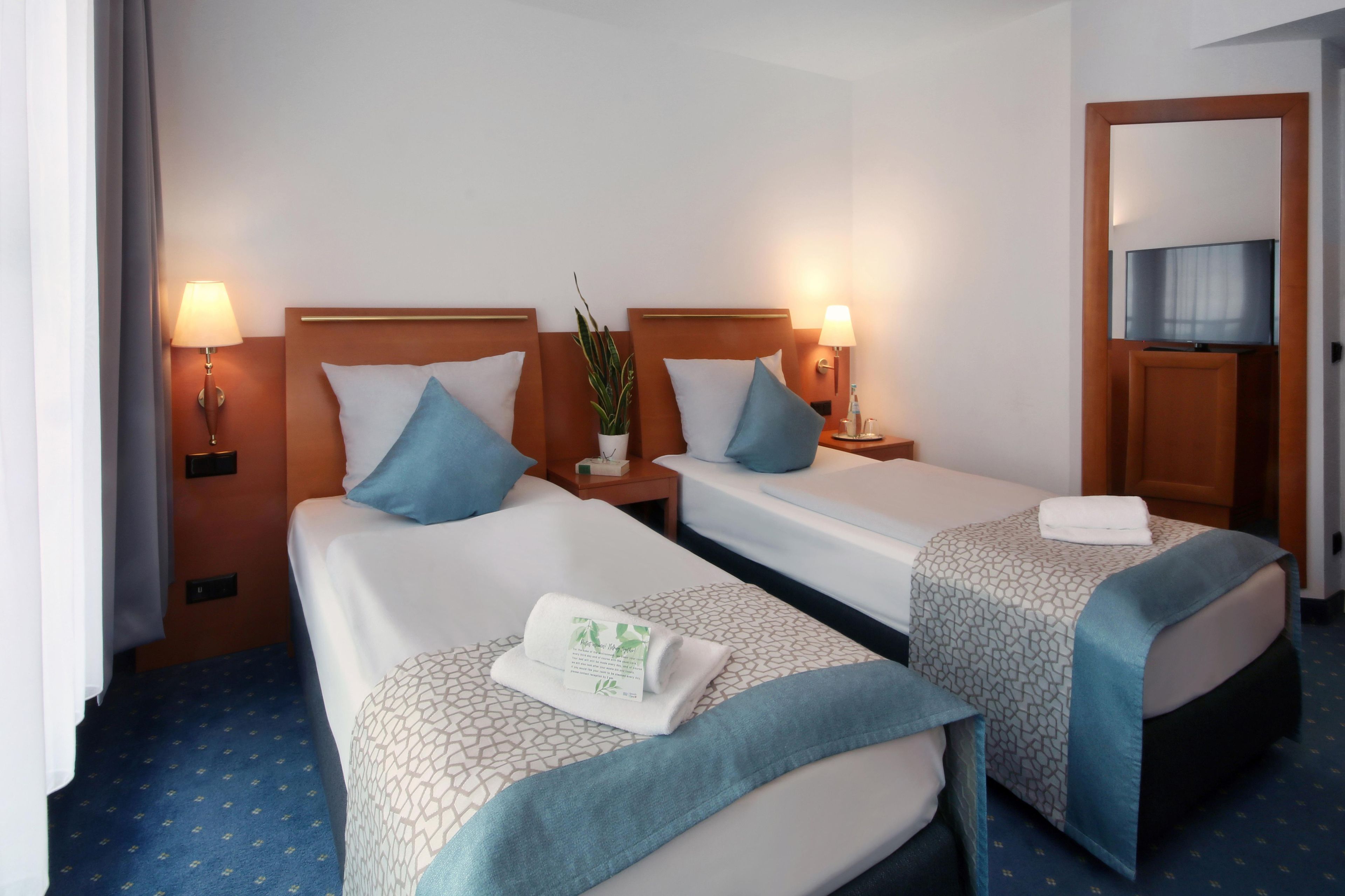 Een hotelkamer met twee bedden, elk met een kussen, een nachtkastje met een lamp en een plant.