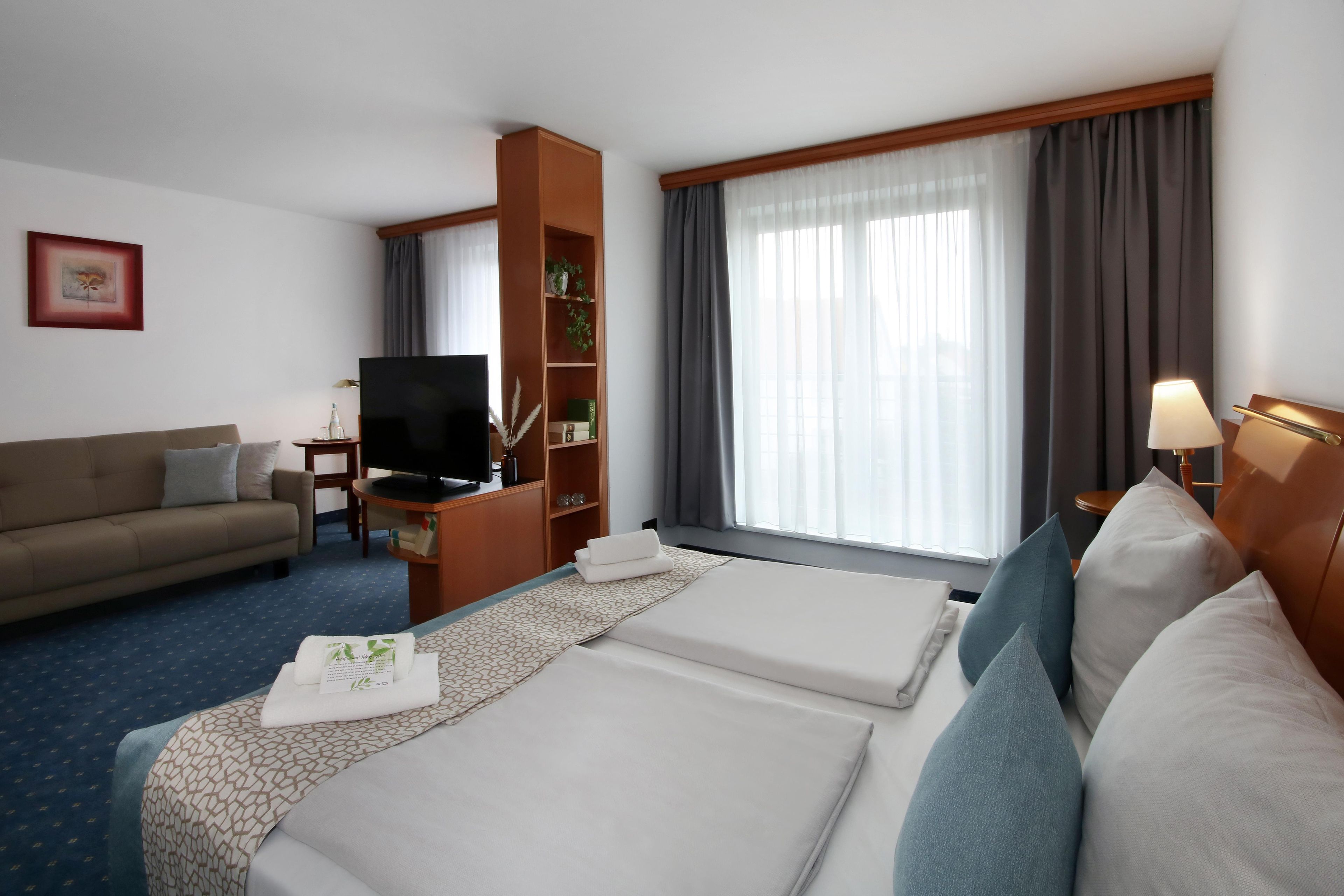 Una habitación de hotel con cama, sofá, televisor, estantería y ventana con cortinas.