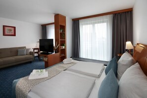 Una habitación de hotel con cama, sofá, televisor, estantería y ventana con cortinas.