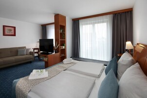 Una habitación de hotel con cama, sofá, televisor, estantería y ventana con cortinas.