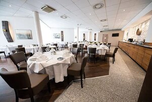 Uma sala de jantar ampla com várias mesas preparadas para hóspedes, com design moderno, toalhas de mesa brancas e cadeiras escuras.
