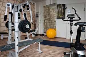 Uma academia com um banco de musculação, uma estrutura para agachamentos, uma prensa de supino, uma bicicleta ergométrica e uma bola de exercício amarela.