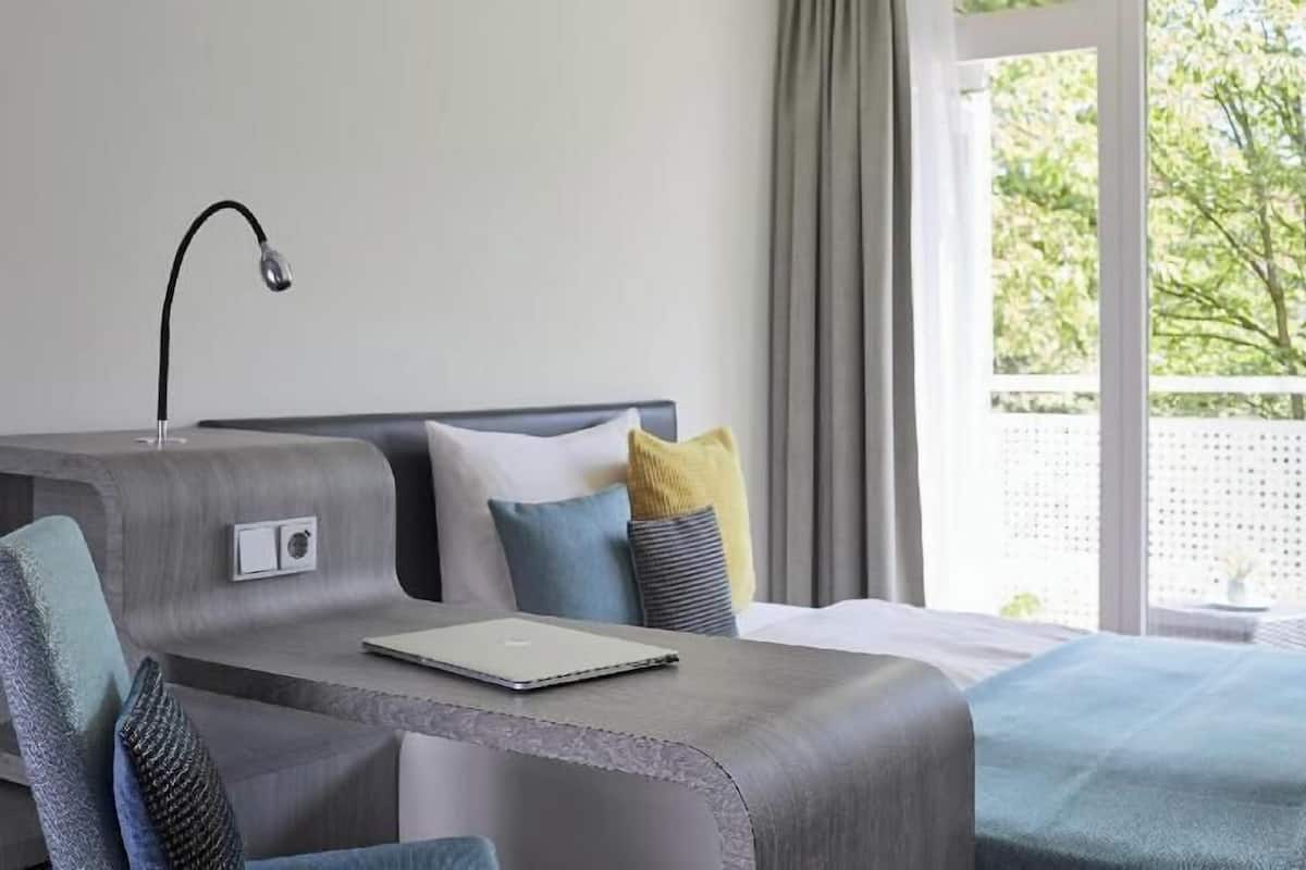 A hotel room with a bed, a desk with a laptop, a chair, and a window with a view of greenery.
